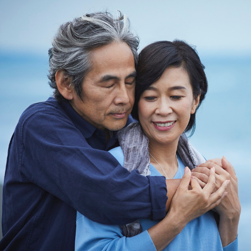 An older couple standing by the water, the man hugging the woman from behind as they both smile