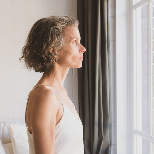 An older woman with wavy gray hair in a cream tank top gazing thoughtfully out of a bright window