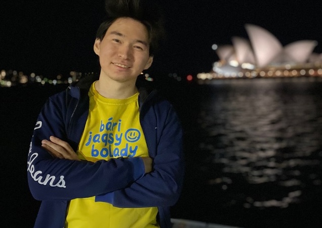 Anuar Aimoldin standing at night in Sydney with the Opera House visible in the background, wearing a yellow t-shirt with 'bári jaqsy bolady' text and a navy blue jacket