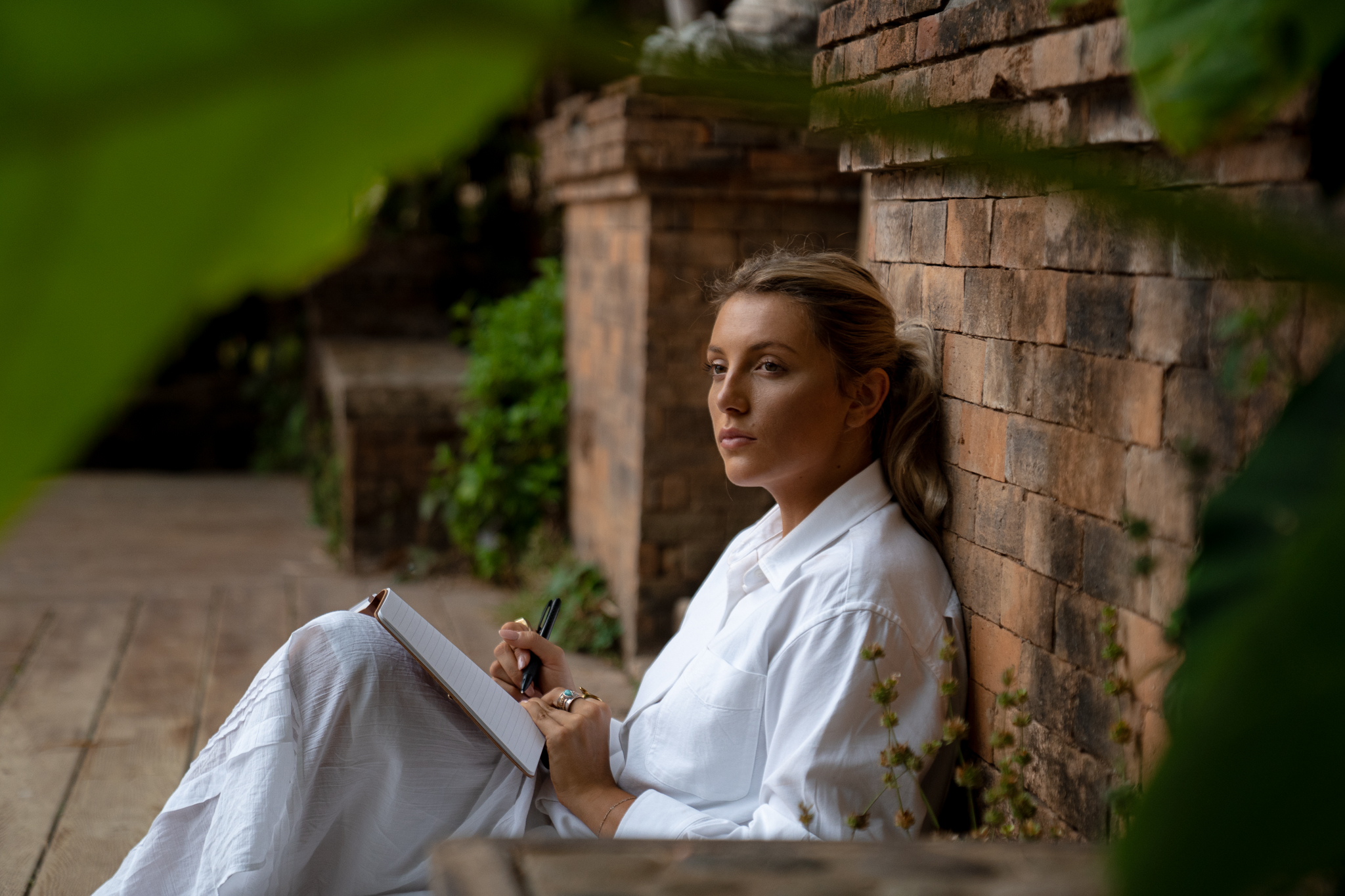 Anna in white linen journaling against brick wall surrounded by tropical plants
