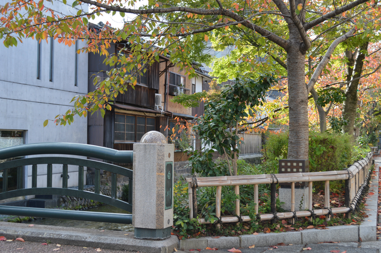 Shirakawa canal in autumn with green arched bridge, stone pillar marker, bamboo fence, and colorful fall foliage framing traditional machiya buildings