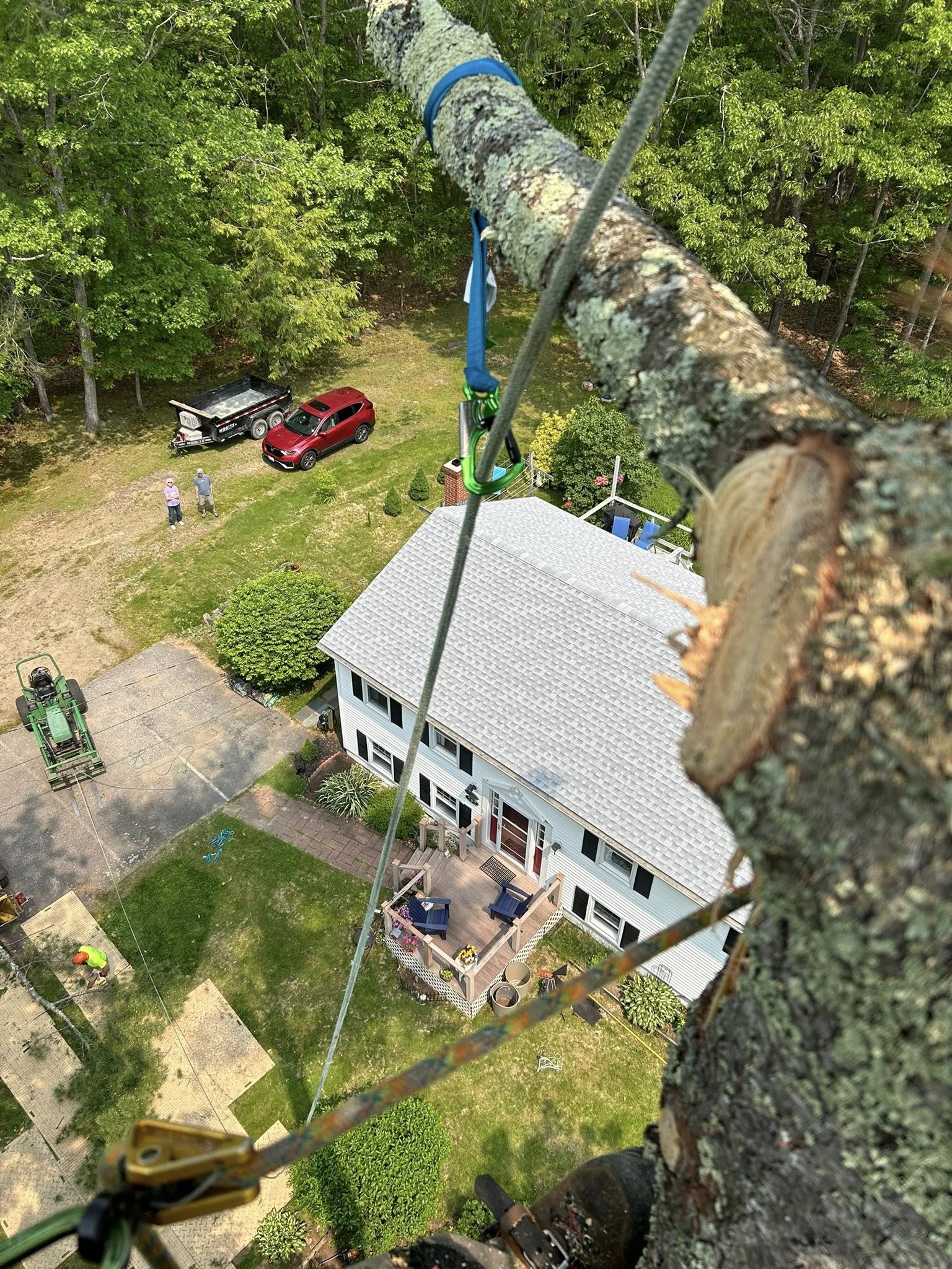 Close-up aerial POV of a large rigged log section suspended mid-air on a carabiner above a residential home and green lawn far below
