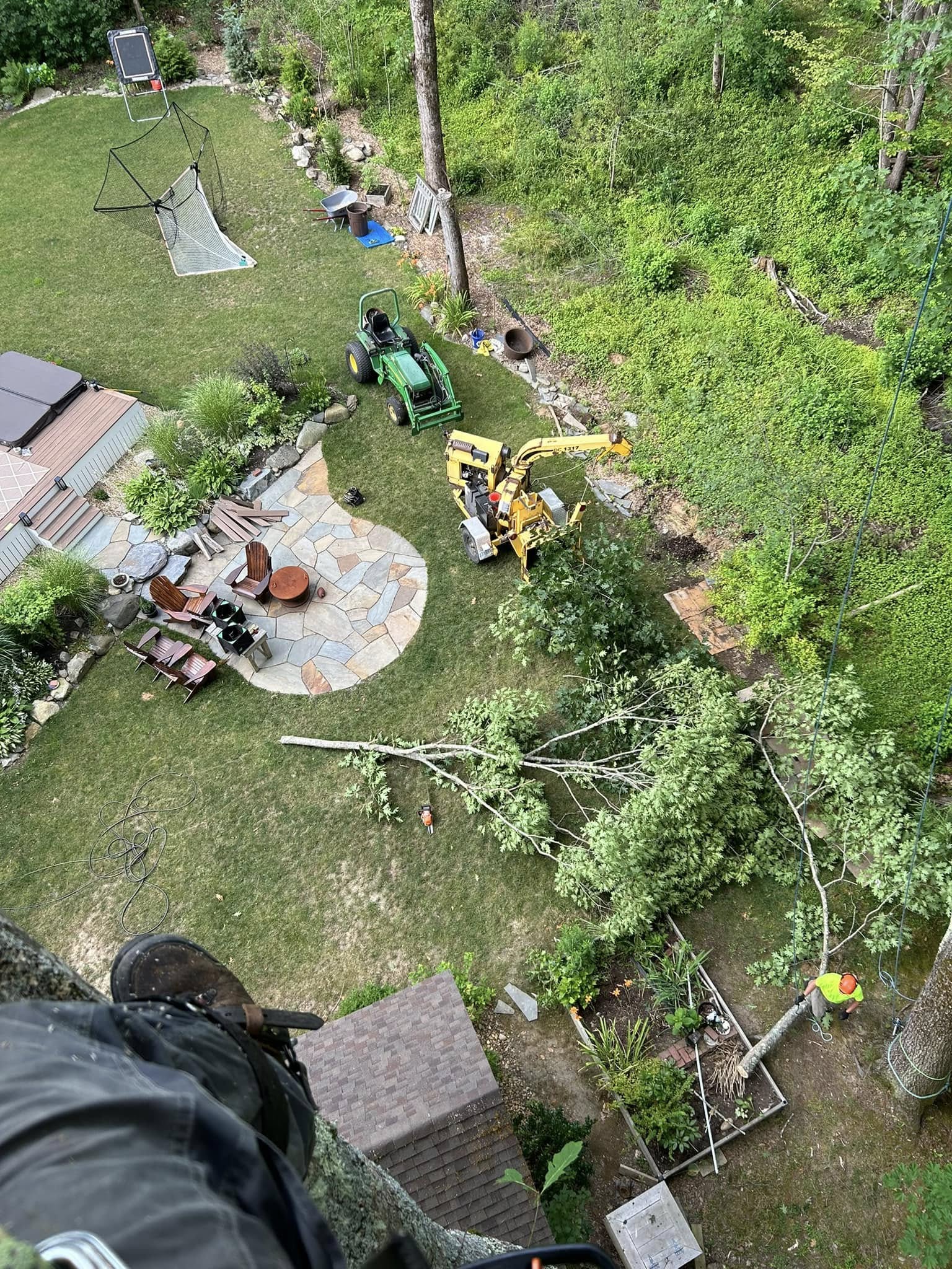 Climber POV looking down at a backyard stone patio with crew in hi-vis, John Deere tractor, chipper, and felled branches on the lawn