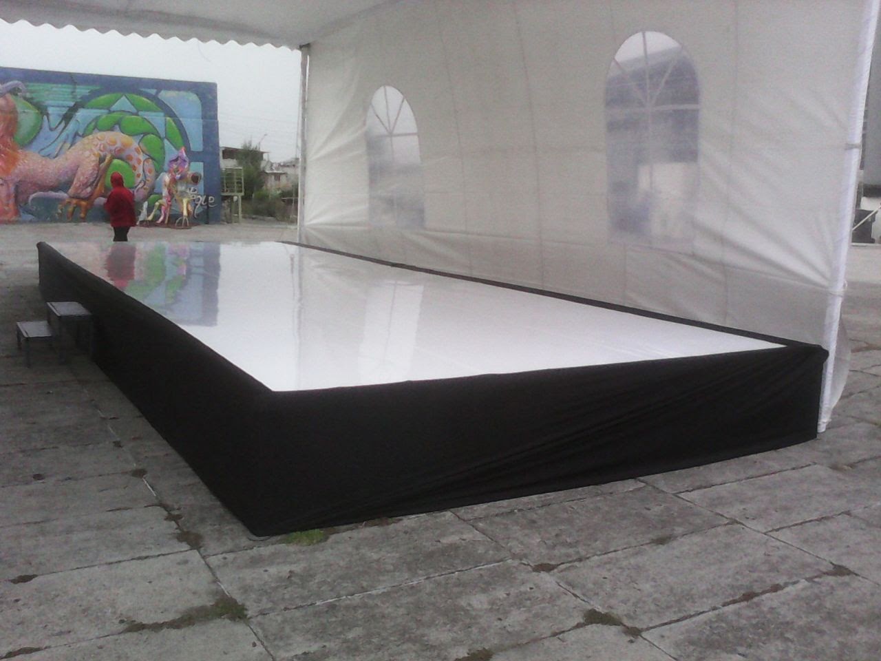 Tarima blanca con falda negra bajo carpa en espacio exterior con mural colorido de fondo