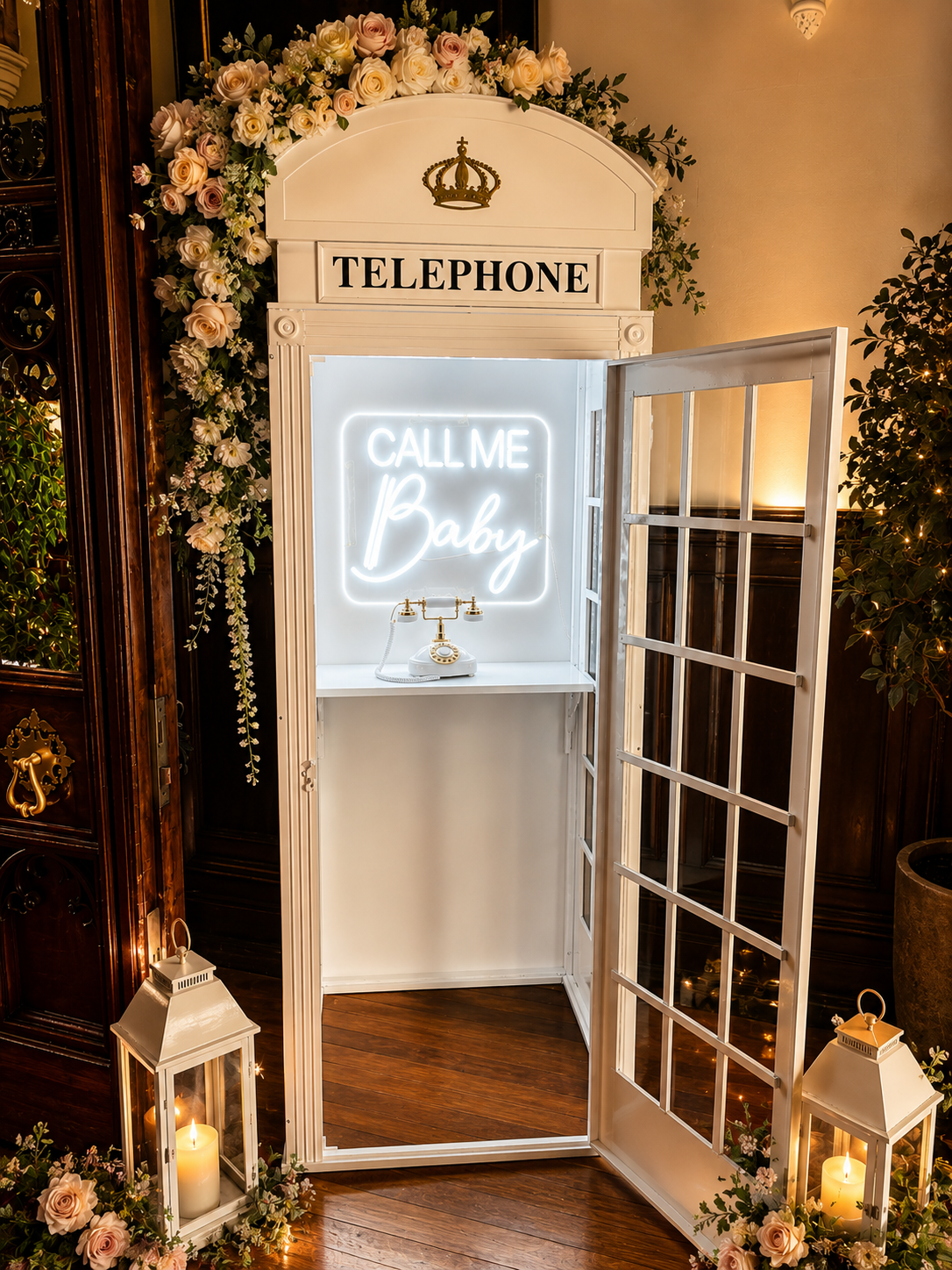 Illuminated white telephone box with Call Me Baby neon sign and antique rotary phone surrounded by roses