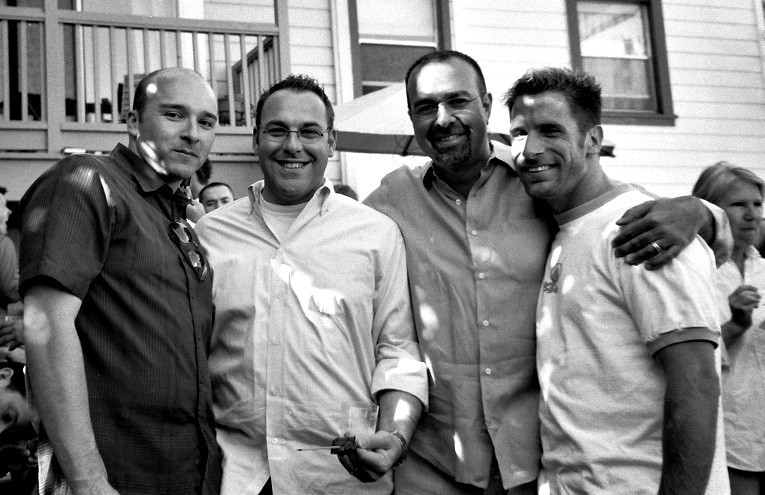 Four men posing together outdoors at gathering with arms around each other in casual camaraderie