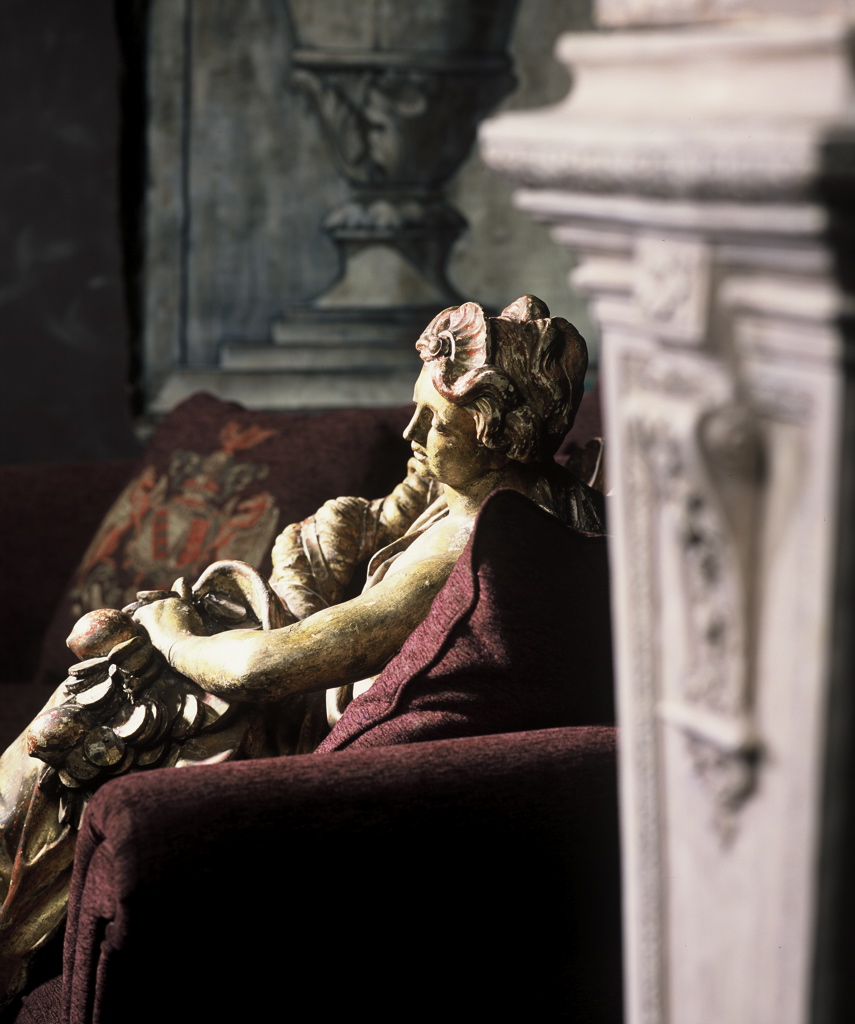 A hand-restored polychrome figure resting against rich burgundy upholstery, framed by trompe l'oeil architecture.