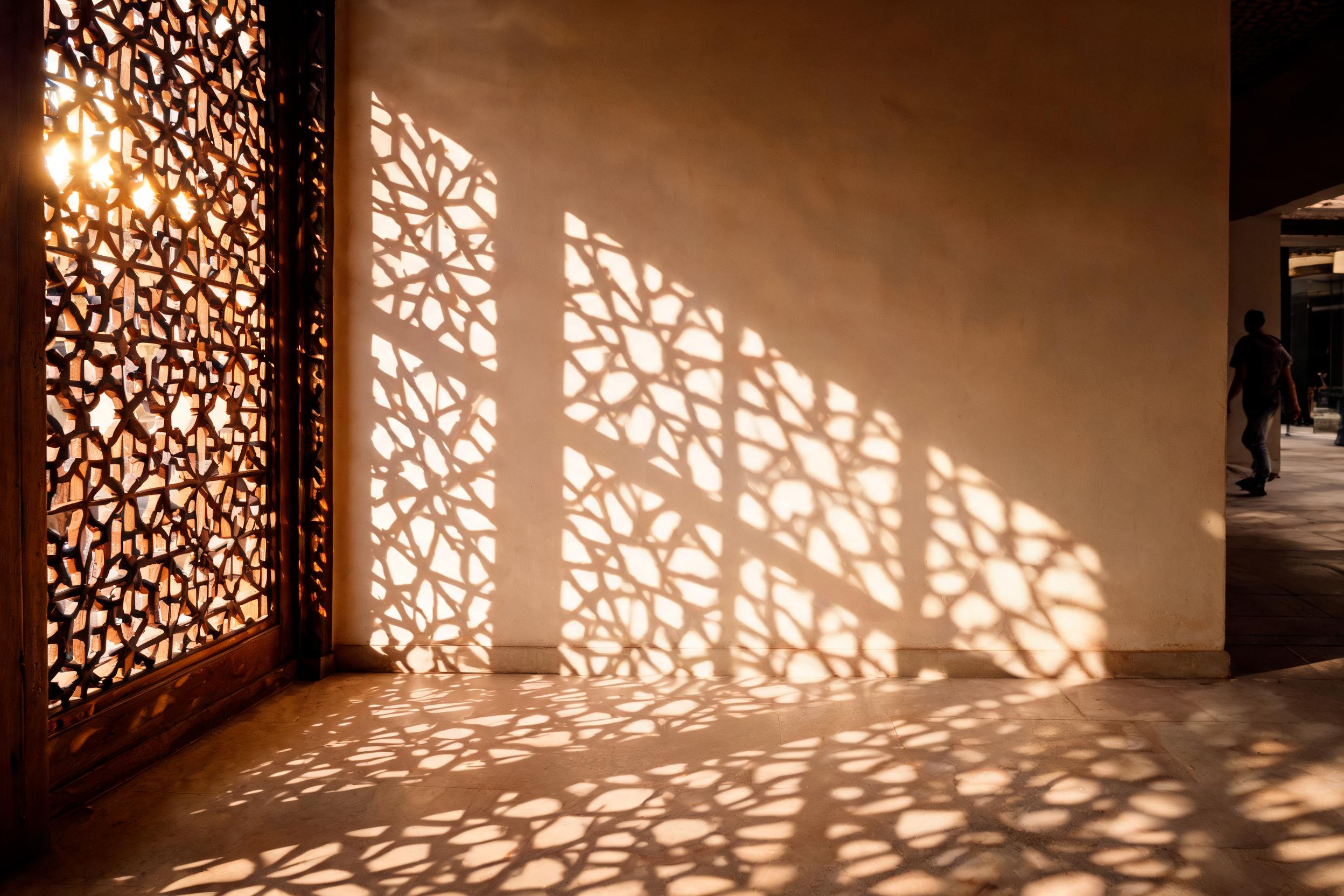 Soft daylight falling through a carved mashrabiya screen, casting interlaced geometric shadows across a warm plaster wall
