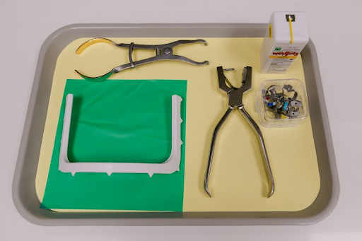 Amalgam tray setup with rubber dam clamp forceps, green rubber dam on frame, hole punch, and matrix clamps