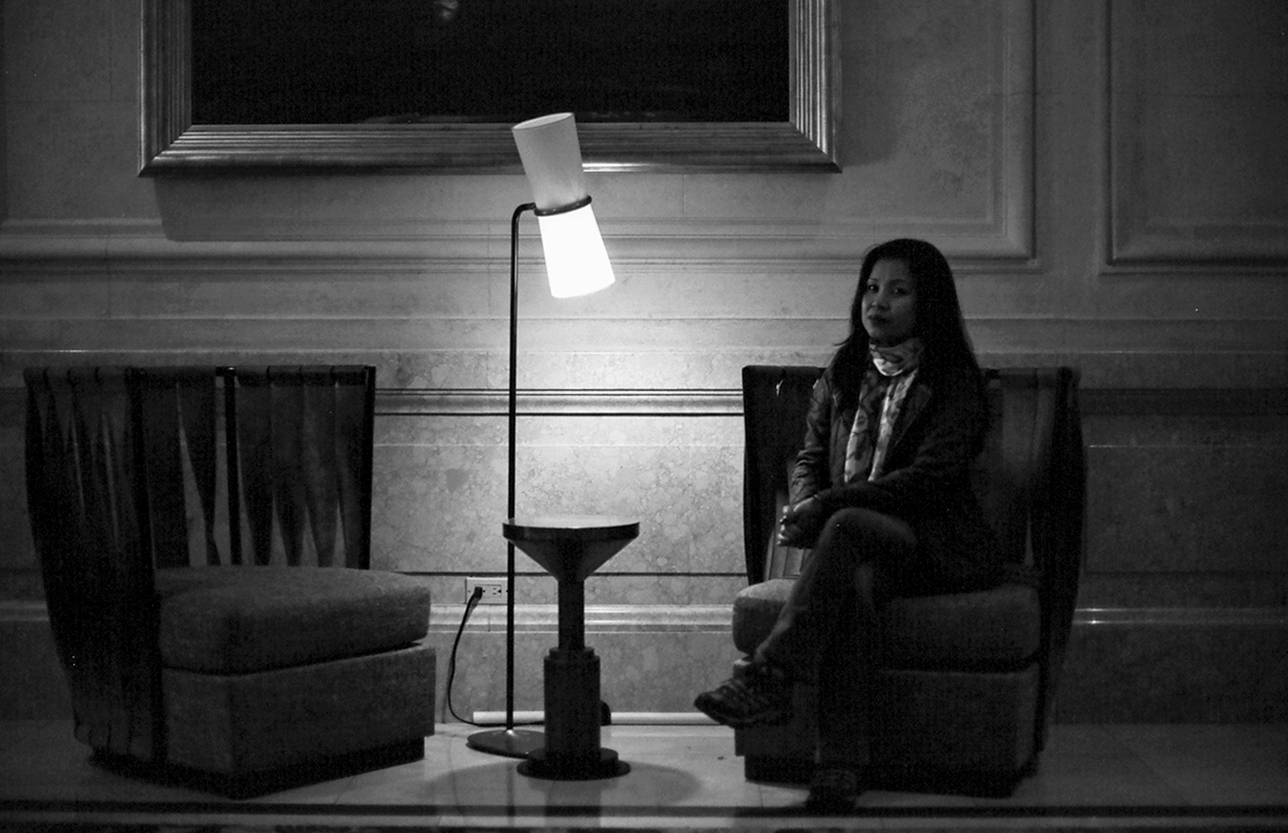 Woman seated in armchair under illuminated desk lamp in minimalist brutalist interior with concrete walls
