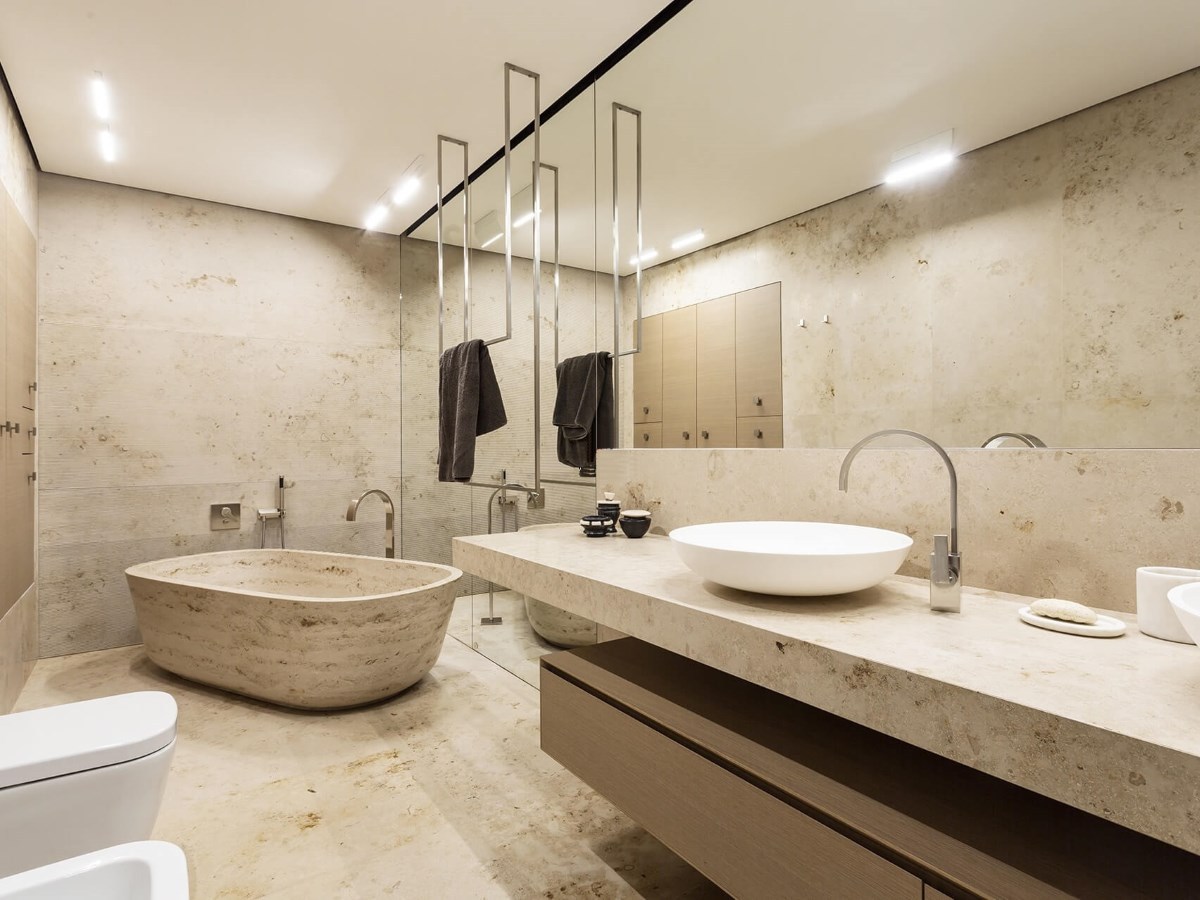 Warm travertine stone bathroom with sculptural stone freestanding tub, vessel sink on stone countertop, stainless mirror frame strips, recessed lighting
