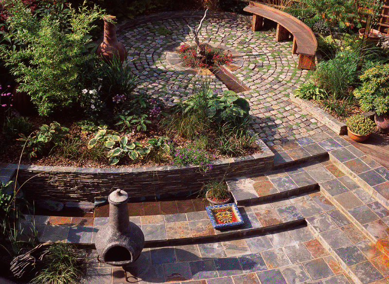 Circular cobblestone patio with a curved wooden bench and lush mixed planting