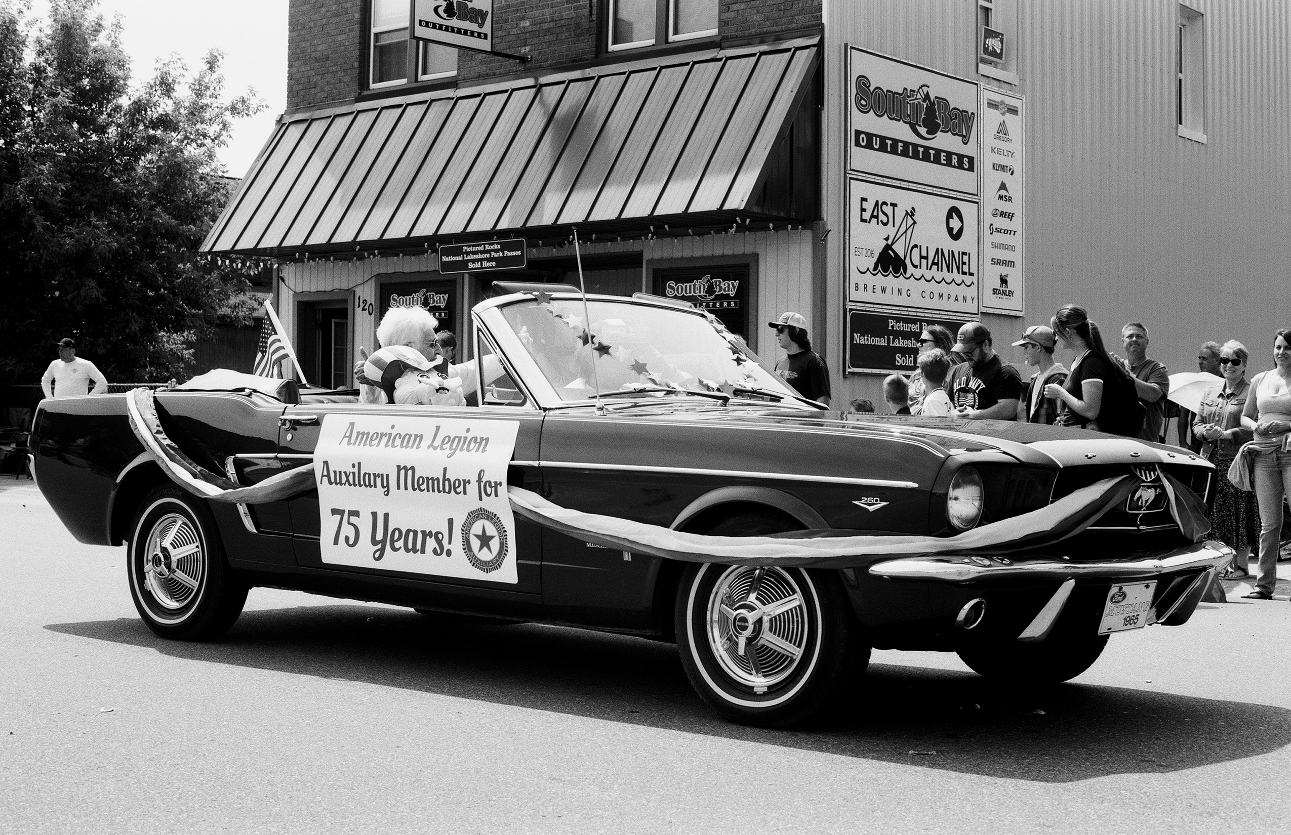 Classic white Mustang convertible in parade with spectators gathered around storefront