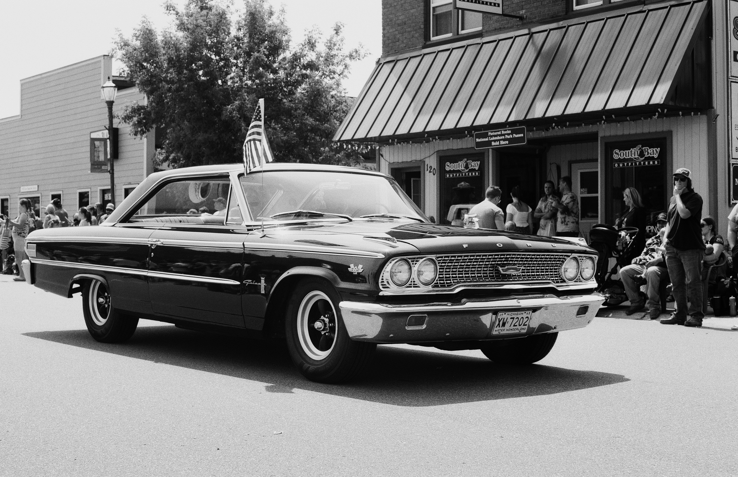 1960s Ford Fairlane parade car with American flag and spectators along storefront