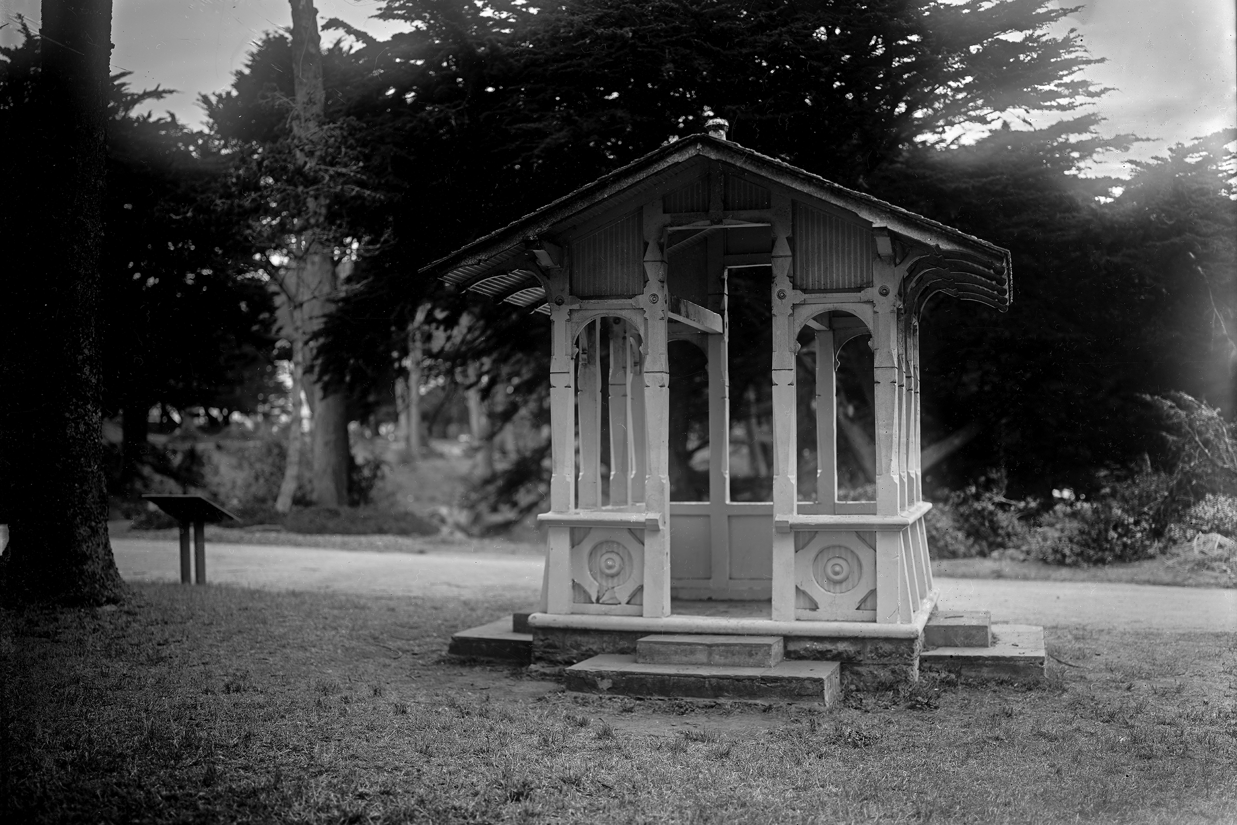 Elegant Victorian glass pavilion kiosk structure in manicured garden with mature trees and lush foliage creating serene composition
