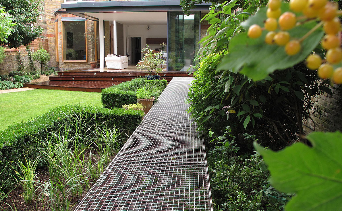 Steel mesh walkway alongside box hedging toward a glass-walled living room