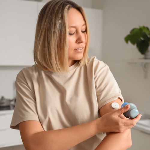 A woman in a kitchen applying a continuous glucose monitor sensor to her upper arm