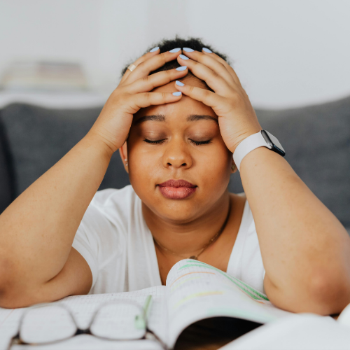 An exhausted woman with her eyes closed and both hands on her head, sitting over an open book and a pair of glasses