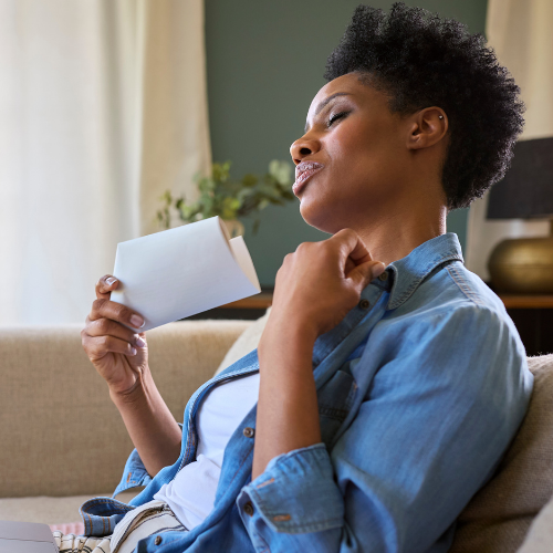 A woman in a denim shirt relaxing on a couch and fanning herself with a folded paper during a hot flash
