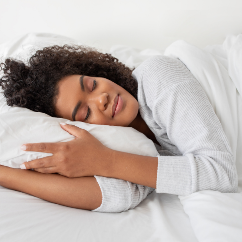 A relaxed woman lying in bed hugging a white pillow, smiling with her eyes closed