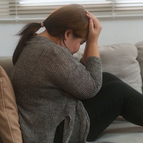 A tired woman in a gray sweater sitting on a couch with her head resting in her hand