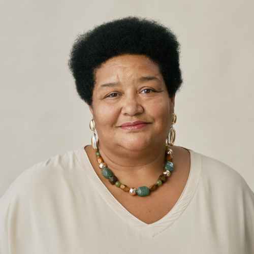 A smiling woman with hoop earrings and a beaded necklace wearing a cream top against a cream background