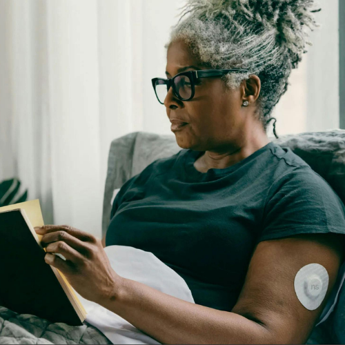 A woman with silver hair and glasses relaxing while reading a book, with a glucose monitor patch on her upper arm