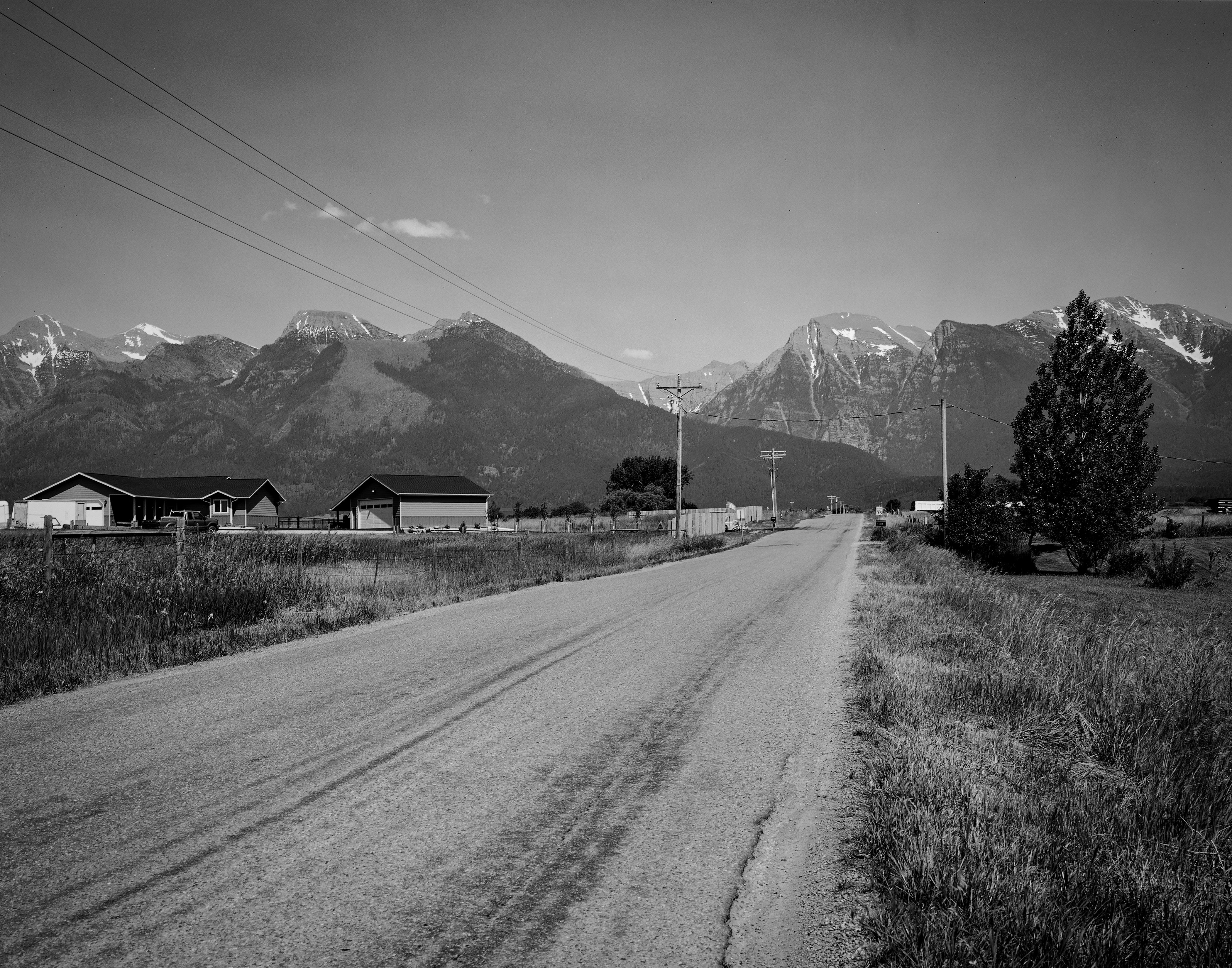 Rural road leading toward snow-capped mountains with farmhouses and power lines under big sky