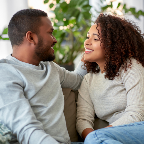 A young couple in cozy sweaters sitting close together on a couch, smiling at each other